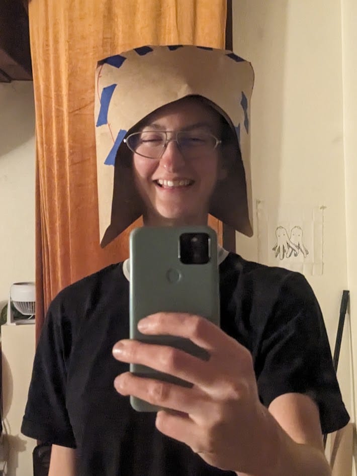 me taking a selfie in a paper prototype of the hood that's way too big in the back. it looks like some kind of medieval knight helmet