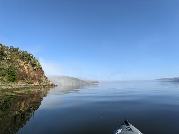 fog burning off over the water, a hilly coastline, flat water, and the front tip of a kayak